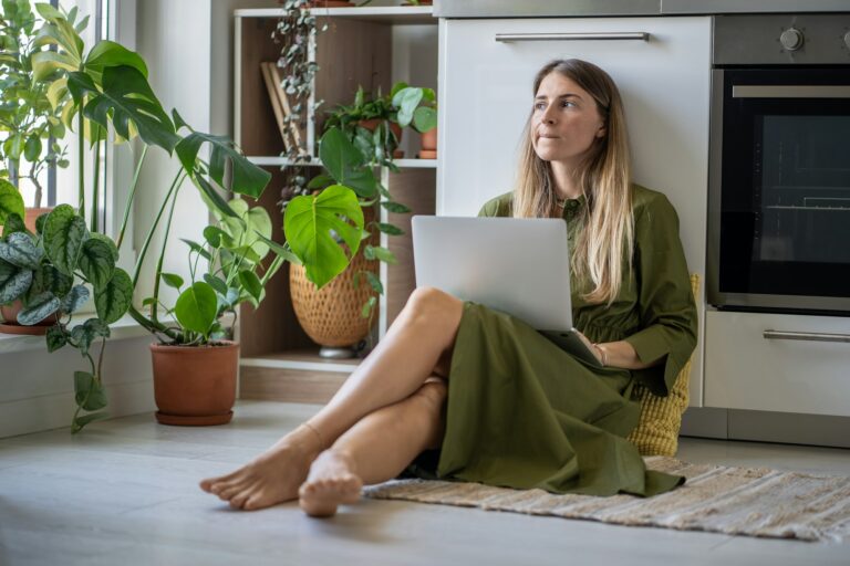 Pensive woman with laptop looking at window, feeling lack of energy sit on floor at home with plants