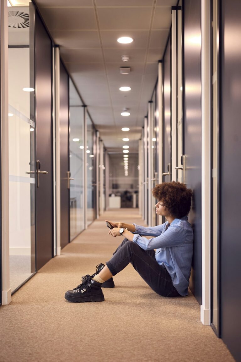 Stressed Young Businesswoman Sitting On Floor In Corridor Of Modern Office With Phone